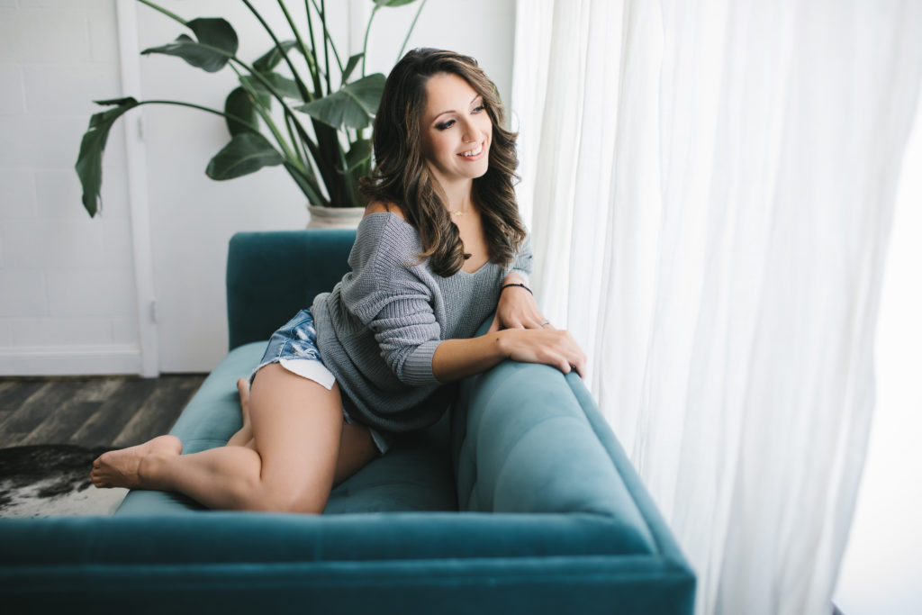 Relaxed Caucasian woman on teal couch looking out a window with white curtains with greenery in the background.  Photography by Lindsay Hite. 