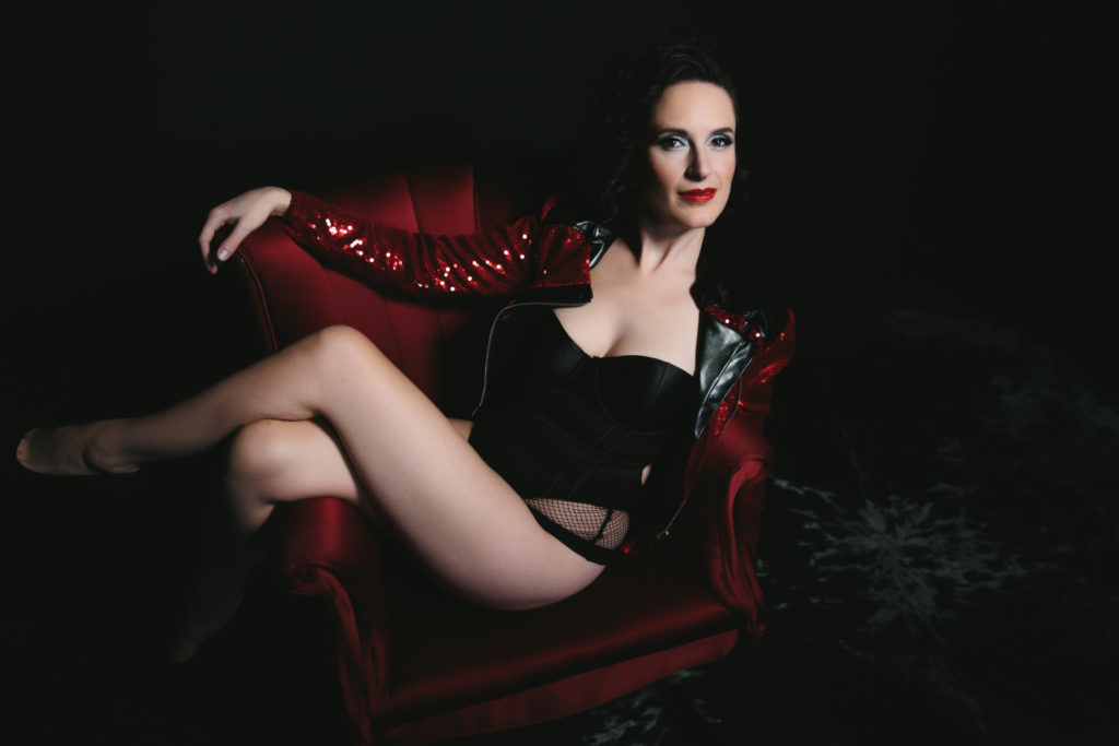 Woman in velvet corset and sparkly red jacket on a red chair with black background.  Photography by Lindsay Hite