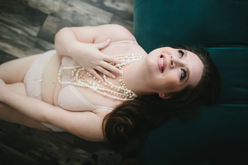 Brunette in pale lingerie and pearl necklaces sitting on a wooden floor leaning against a teal sofa; boudoir photography by Lindsay Hite