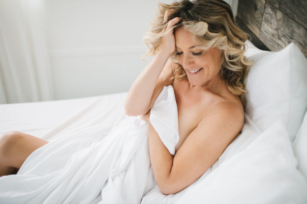 Blonde curly haired woman in white bed sheets on bed with white background.  Photography by Lindsay Hite. 