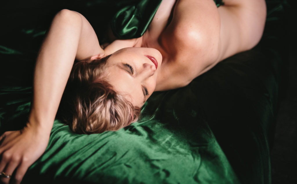Short haired blonde woman in emerald green satin sheets with dark background.  Photography by Lindsay Hite. 