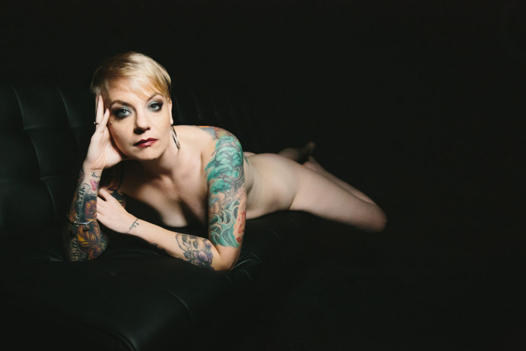 Woman with tattoos in the nude on a black sofa.  Photography by Lindsay Hite