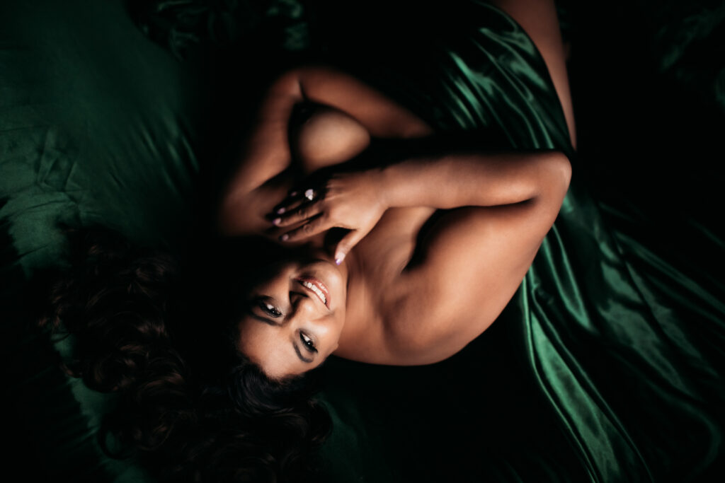 Woman between emerald satin sheets against a black backdrop; boudoir photography by Lindsay Hite