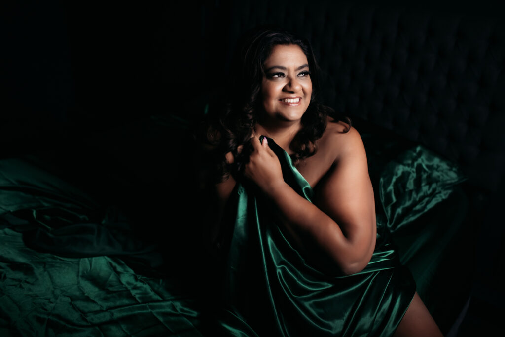 Woman between emerald satin sheets against a black backdrop; boudoir photography by Lindsay Hite