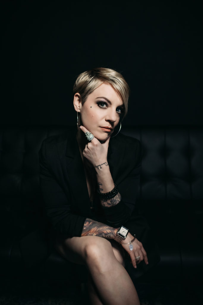 Woman in business suit in seated power pose, with tattoos and jewelry on.  Photography by Lindsay Hite