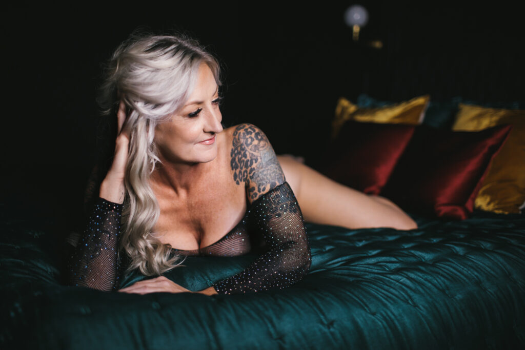 woman over 40 in rhinestone studded mesh bodysuit on a teal velvet bed with a dark backdrop.  Boudoir photography by Lindsay Hite
