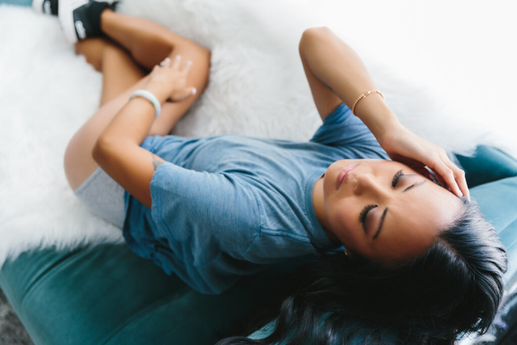 Asian woman in a blue t-shirt and undies with sneakers on leaning back on a teal sofa with a white fur-lined blanket on it.  Photography by Lindsay Hite