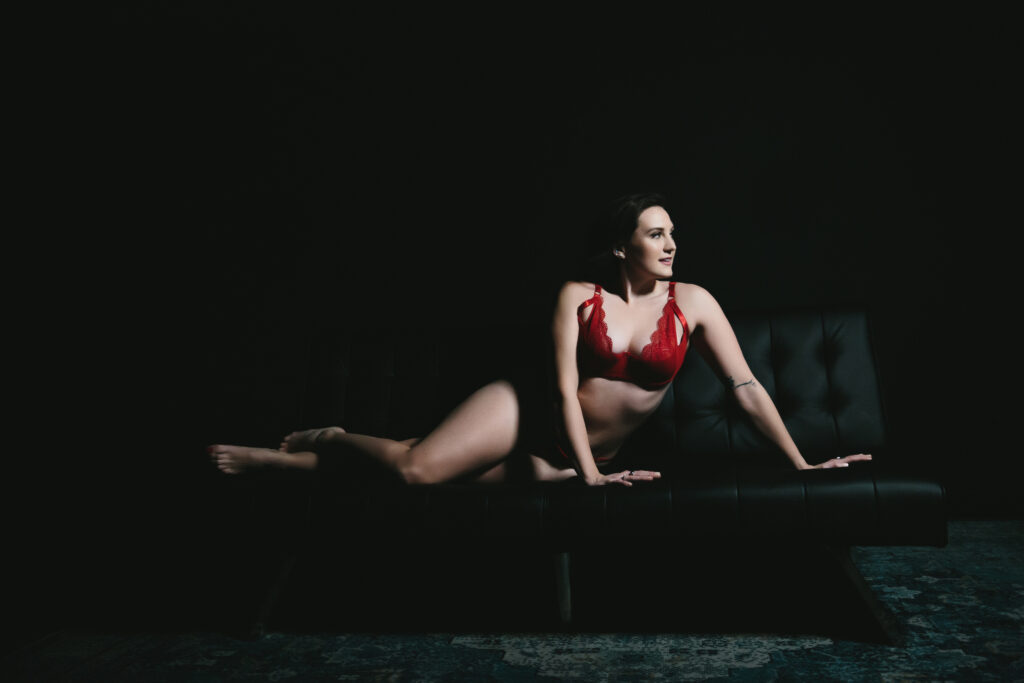 Caucasian woman in red lingerie on a black sofa with a dark background.  Boudoir photography by Lindsay Hite. 