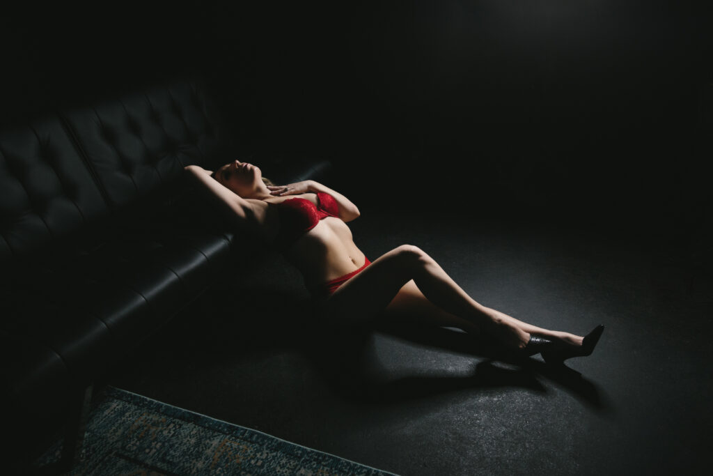 Woman in red lingerie leaning against a black couch with a black floor in a black background.  Photography by Lindsay Hite.