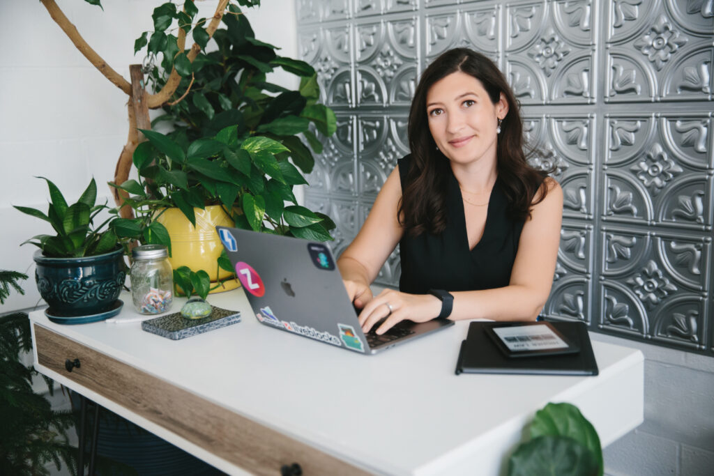 Brunette woman in a black sleeveless top sitting at a desk with her laptop.  Branding photography by Lindsay Hite at Show Your Spark. 