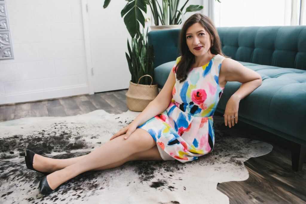Woman in a vibrant colorful floral dress sitting on the floor in front of a teal sofa.  Branding photography by Lindsay Hite at Show Your Spark. 