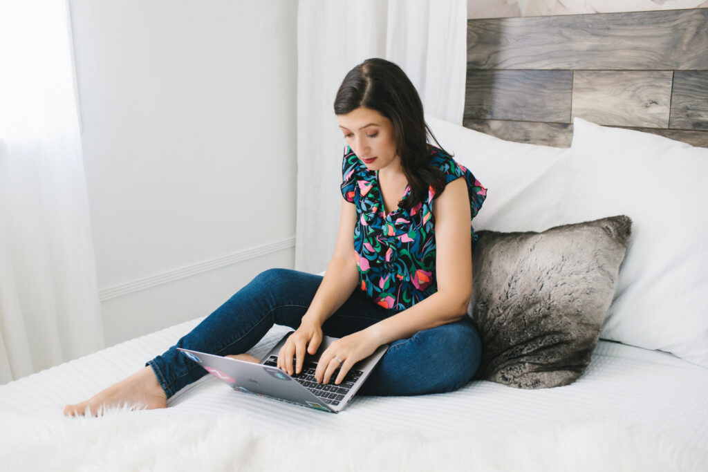 Branding session with a woman sitting on a white bed working on her laptop.  Photography by Lindsay Hite. 