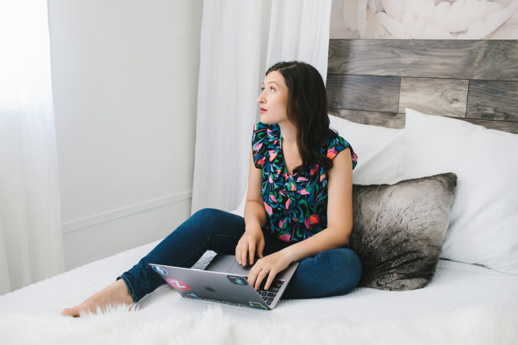Woman wearing a floral top sitting on a white fur-lined bed working on her laptop.  Brunette woman in a red dress with a grey background.  Branding photography by Lindsay Hite at Show Your Spark. 