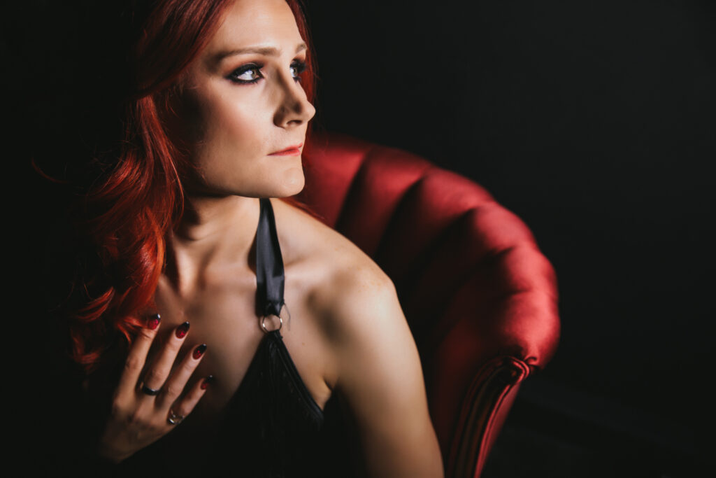 Woman with red hair in a black bra sitting on a red armchair with a black background. Boudoir photography by Lindsay Hite. 