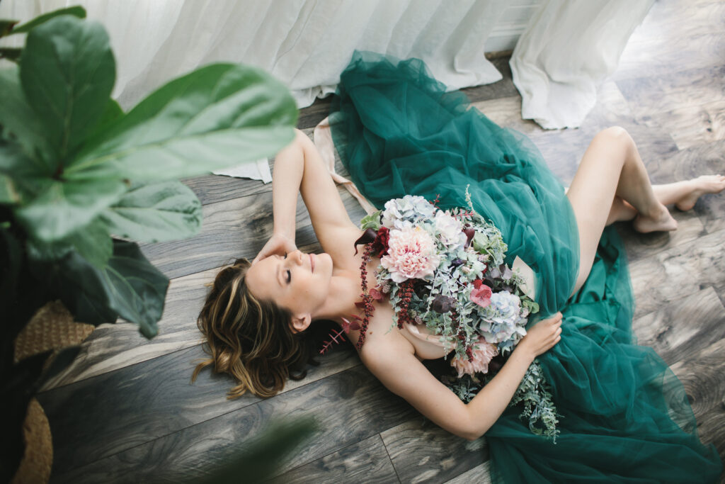 Blonde Caucasian woman in green toile and a floral bouquet laying on the floor. Boudoir photography by Lindsay Hite. 
