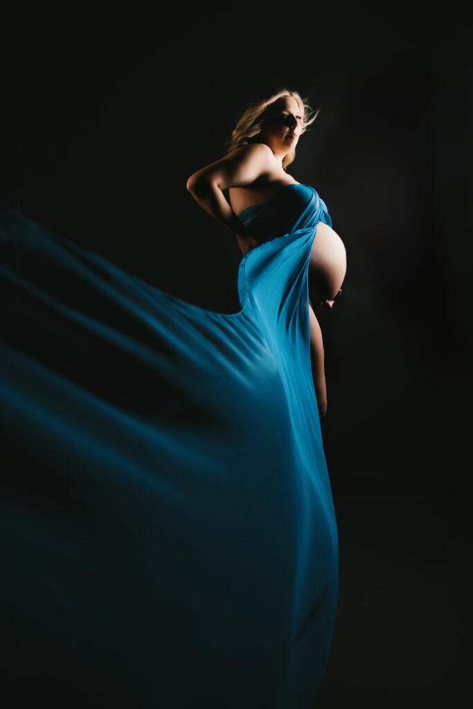 Dramatic image of a pregnant woman in flowing blue silks with a black backdrop and windswept hair.  Photography by Lindsay Hite