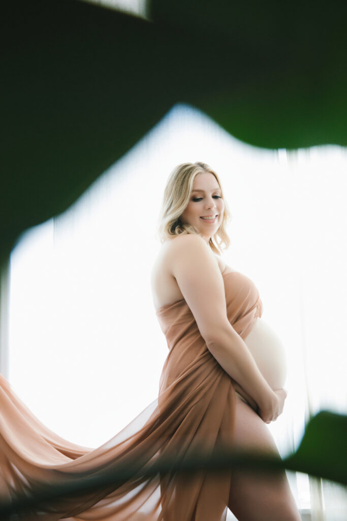 Pregnant woman in beige silks with a white backdrop.  Embrace your pregnant body through maternity boudoir photography.  Photography by Lindsay Hite