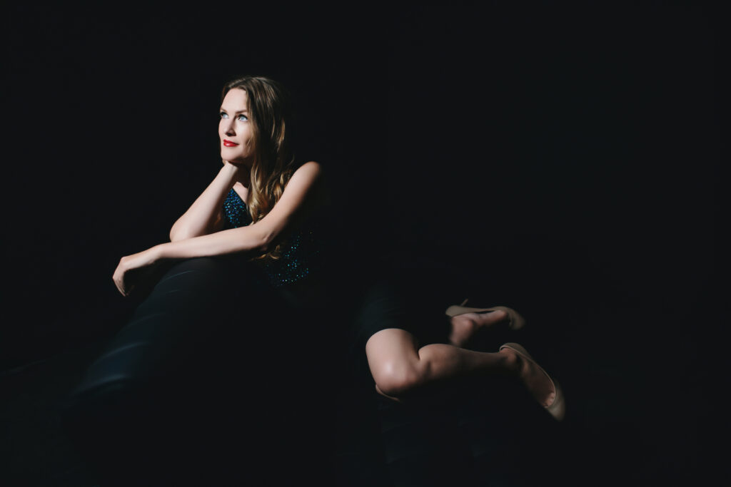 woman in green dress sitting on a black sofa with a black background.  Moms Need a Boudoir Session.  Photography by Lindsay Hite