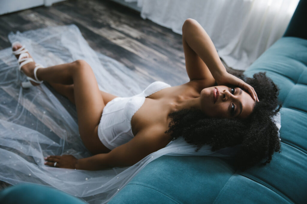 African America woman in a white corset with a long bridal veil sitting on the floor leaning up again a teal sofa. Photography by Lindsay Hite at Show Your Spark.