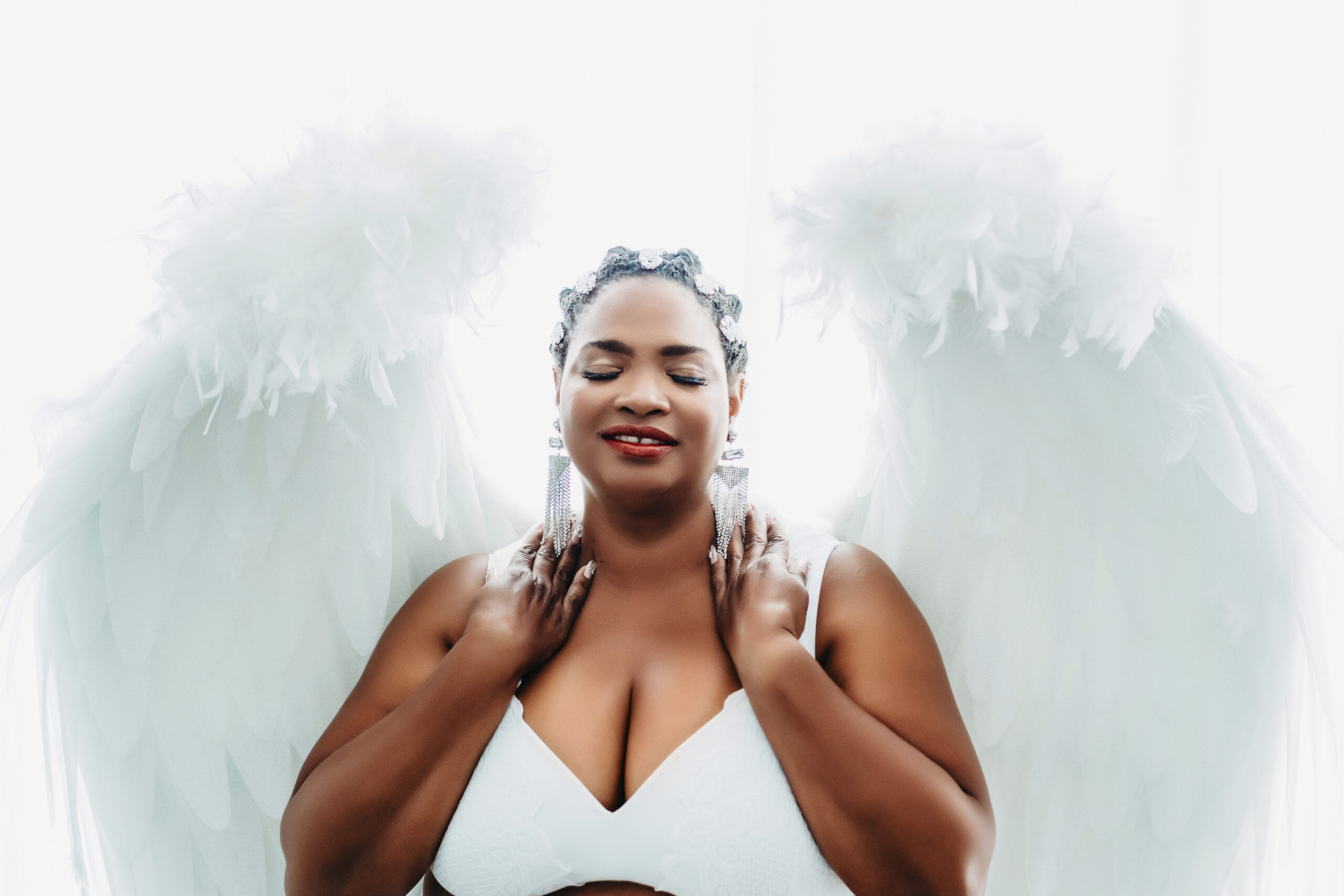 African American woman in a white bra with white angel wings with a white backdrop. Boudoir photography by Lindsay Hite at Show Your Spark.