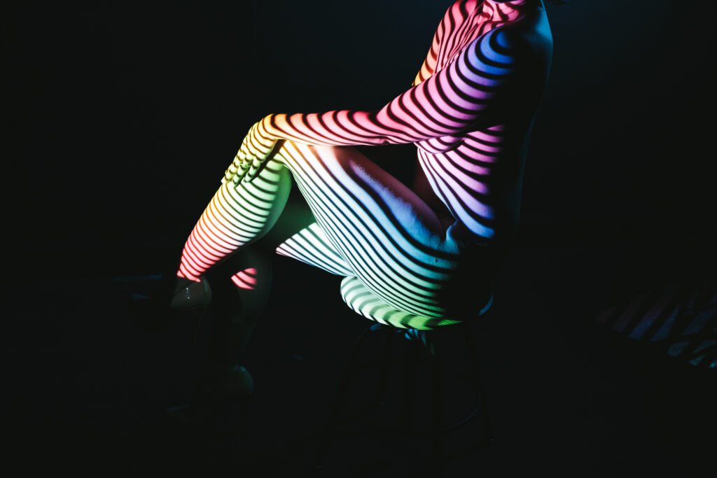 Image of nude woman sitting in a chair with neon stripes superimposed over her with a dark background. Photography by Michelle DeVoe at Show Your Spark