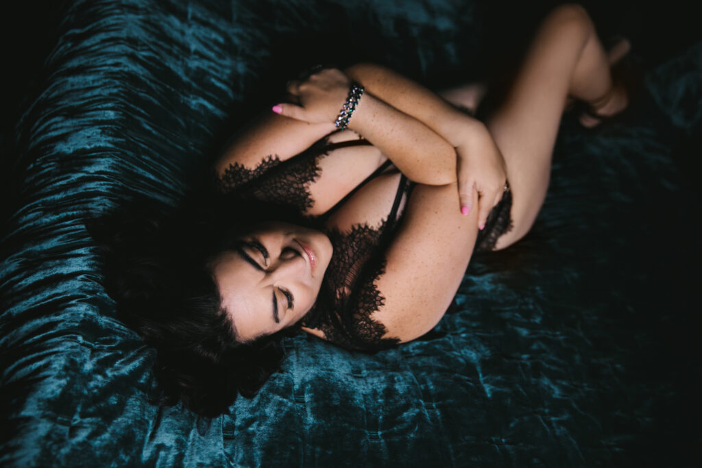 Woman in a black lace body suit in a dark room on her back on a dark teal jewel-toned bed declaring her fierceness.  Photography by Lindsay Hite