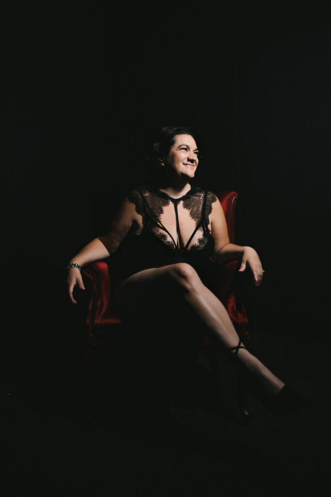 Woman smiling big in a lace bodysuit sitting in a red velvet chair with a black backdrop.  Photography by Lindsay Hite