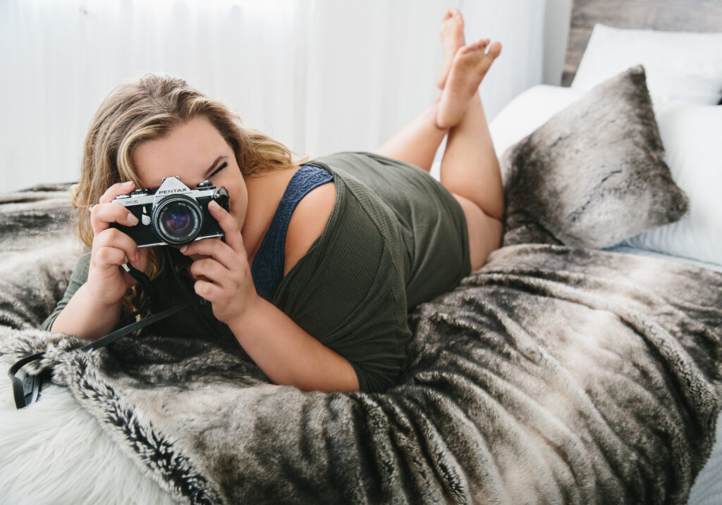 Woman in a t-shirt on her belly with a camera on her face as if she is taking a picture.  Self-care can also involve doing activities that bring you joy.  Photography by Lindsay Hite