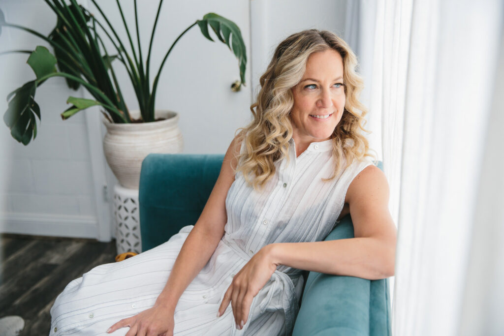 Blonde woman in a white dress sitting on a teal sofa looking out the window through white curtains.  Professional photography services by Lindsay Hite. 