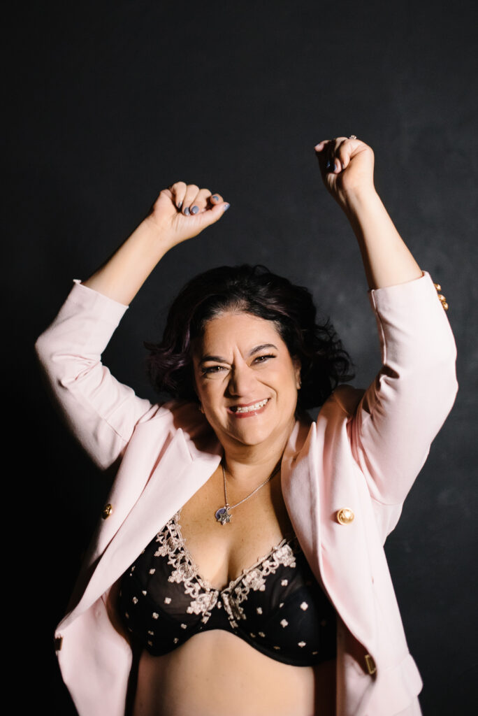 Woman in a black bra and pink jacket smiling with a grey background.  Photography by Lindsay Hite
