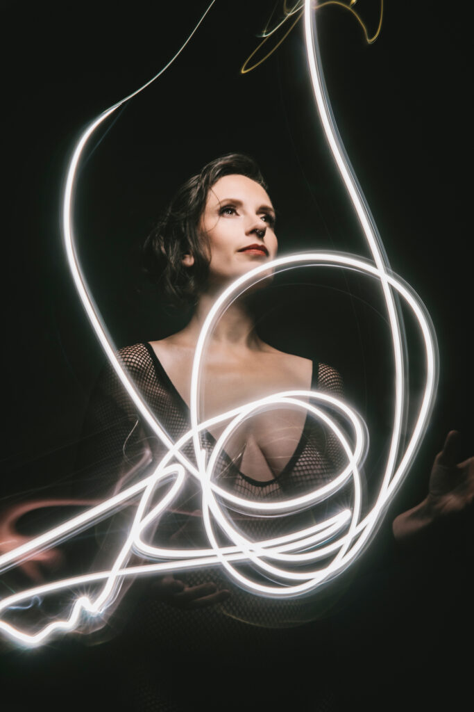 Woman with fishnet bodysuit with a dark background with neon lights in front of her.  Photography by Lindsay Hite. 