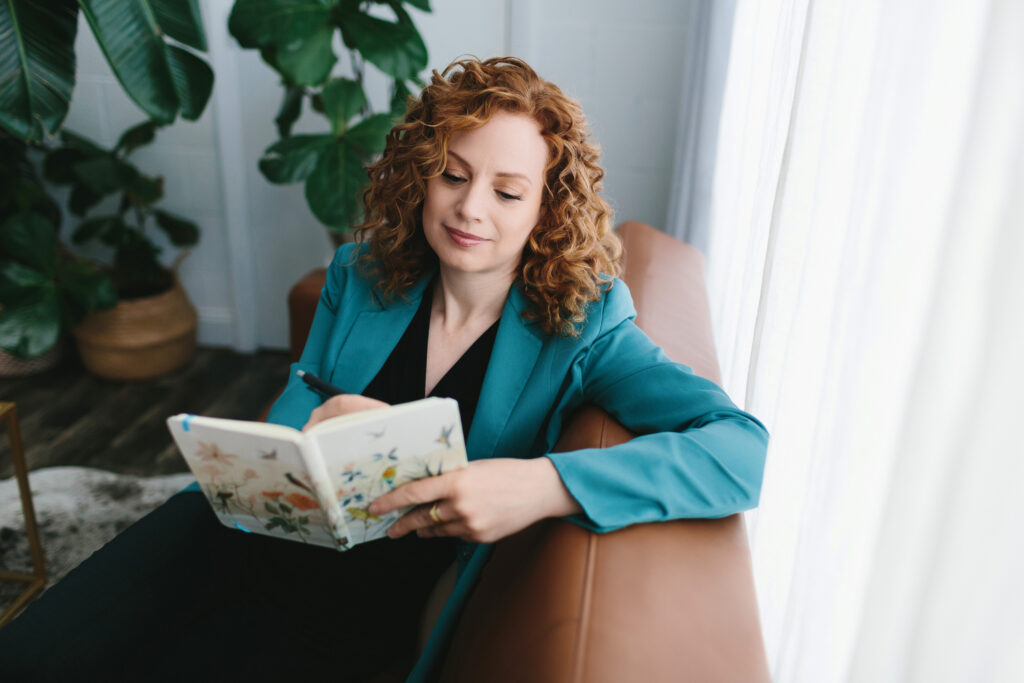 Woman in black blouse and teal jacket sitting on a leather sofa with a journal. writing about what 
she's learned lessons from adversity in our Fierce After Forty project by Lindsay Hite. 