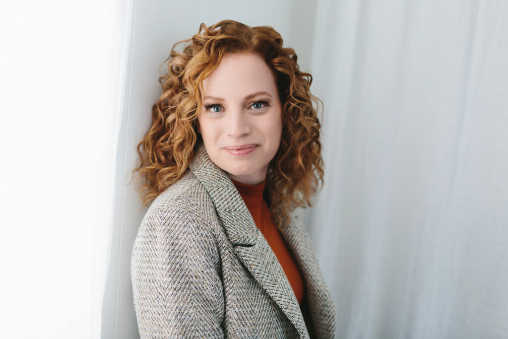 Woman in tweed jacket and orange blouse against a white curtain backdrop.  In this blog post she speaks of lessons from adversity.  Photography by Lindsay Hite