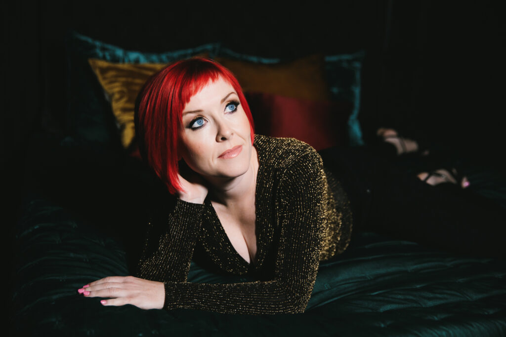 Woman with bright red hair and deep blue eyes in a gold shirt laying on a dark teal bed.  Photography by Lindsay HIte