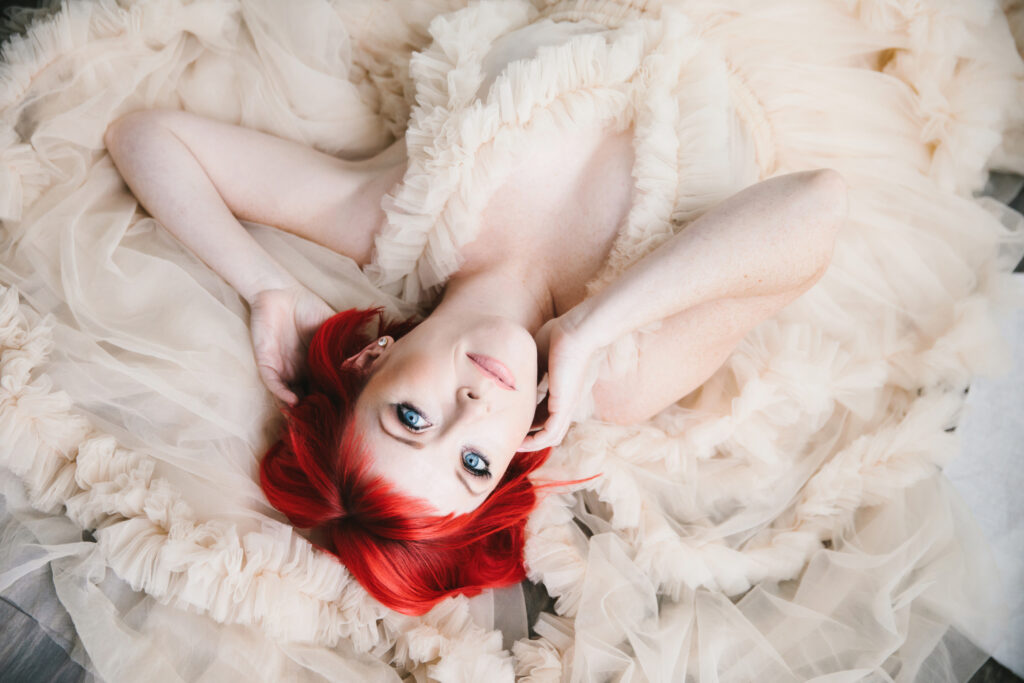 Woman with bright red hair and deep blue eyes in a white fluffy dress laying on the ground.  Boudoir photography by Lindsay Hite. 