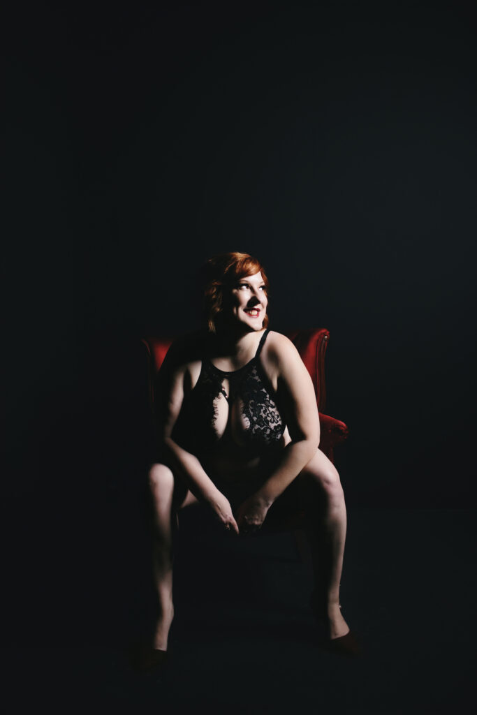 Woman in black lingerie sitting in a red velvet chair with a black backdrop looking into the future while thinking of the most important lesson learned after forty-- prioritize self-care. Photography by Lindsay Hite