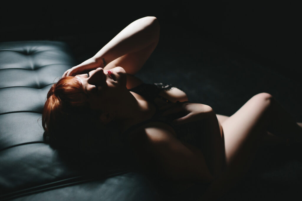 Woman in black lingerie leaning back against black sofa with a dark background. Photography by Lindsay Hite