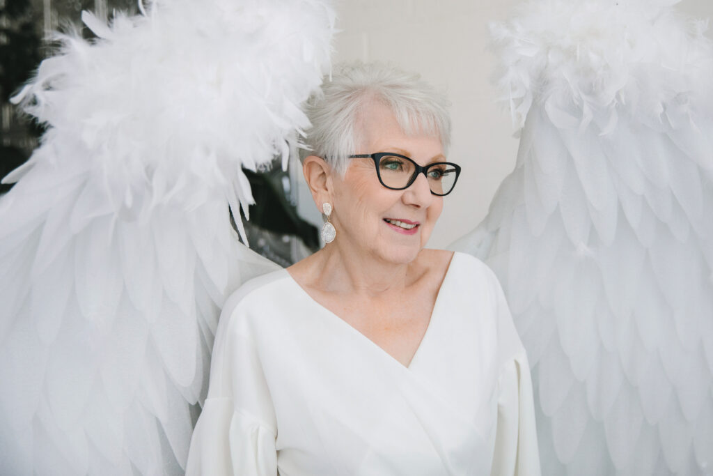 Woman in her 80's in a a white top and angel wings looking in the distance with love in her eyes.  Photography by Lindsay Hite. 