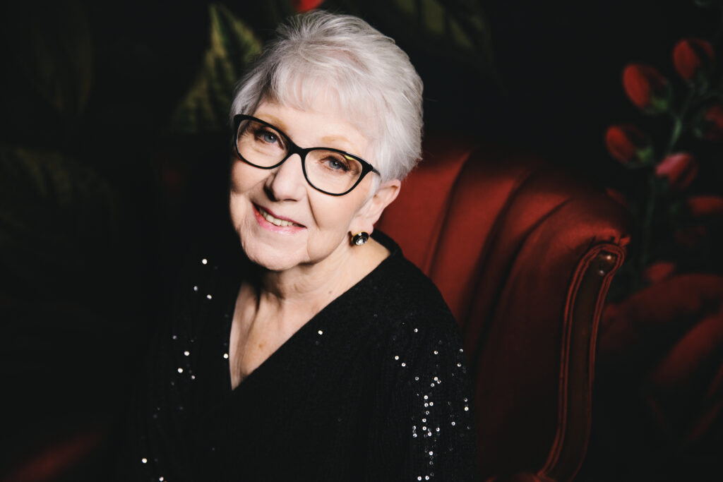 Woman in her 80's in black sparkly top sitting in a red velvet chair with floral backdrop showing off her fierce spirit.  Photography by Lindsay Hite. 