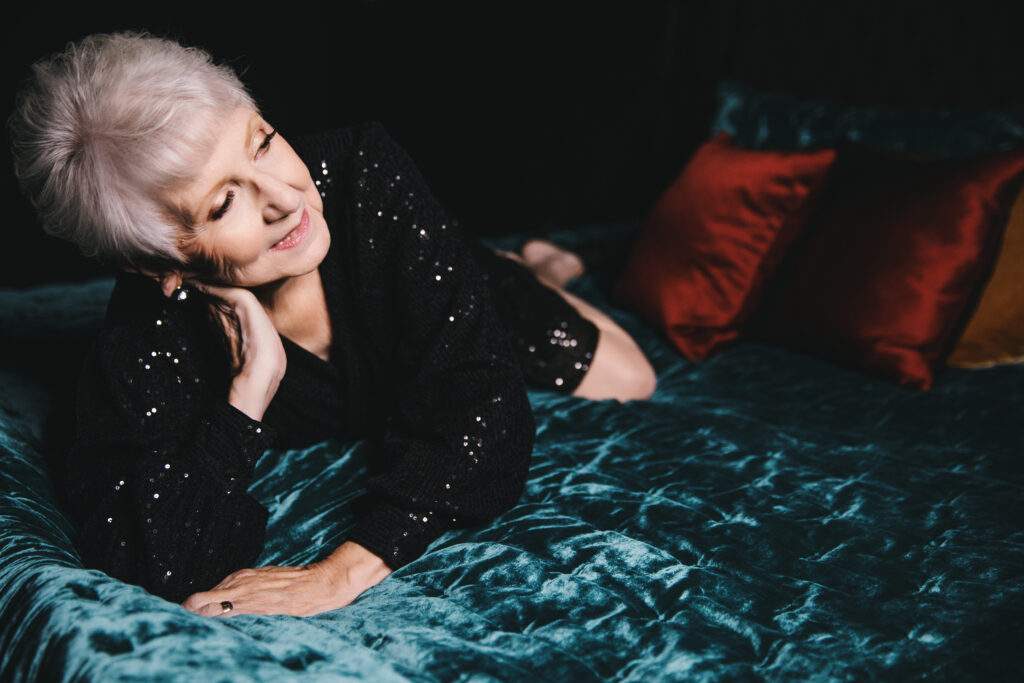 Woman in her 80's in sparkly black top on a green jewel-toned bed.  Photography by Lindsay Hite. 