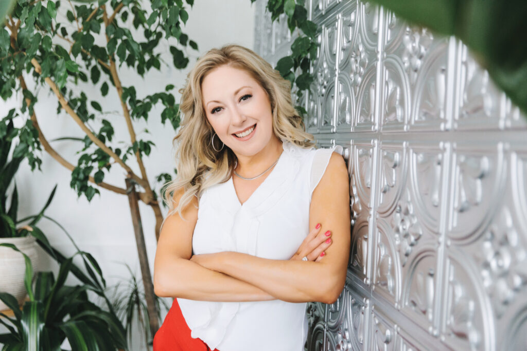 Woman standing in power pose, leaning against a silver wall in a white shirt and red pants.  Branding session portraits by Lindsay Hite. 