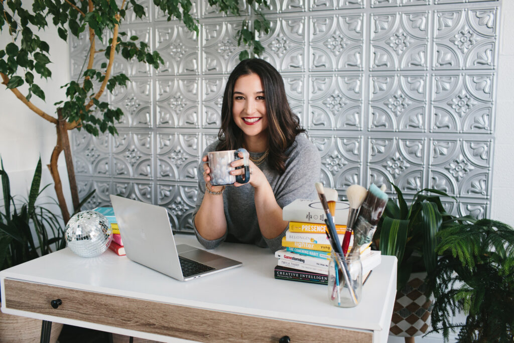 Branding photography session with a woman sitting at a desk with a computer, coffee mug, books, and painting supplies.  Photography by Lindsay Hite. 