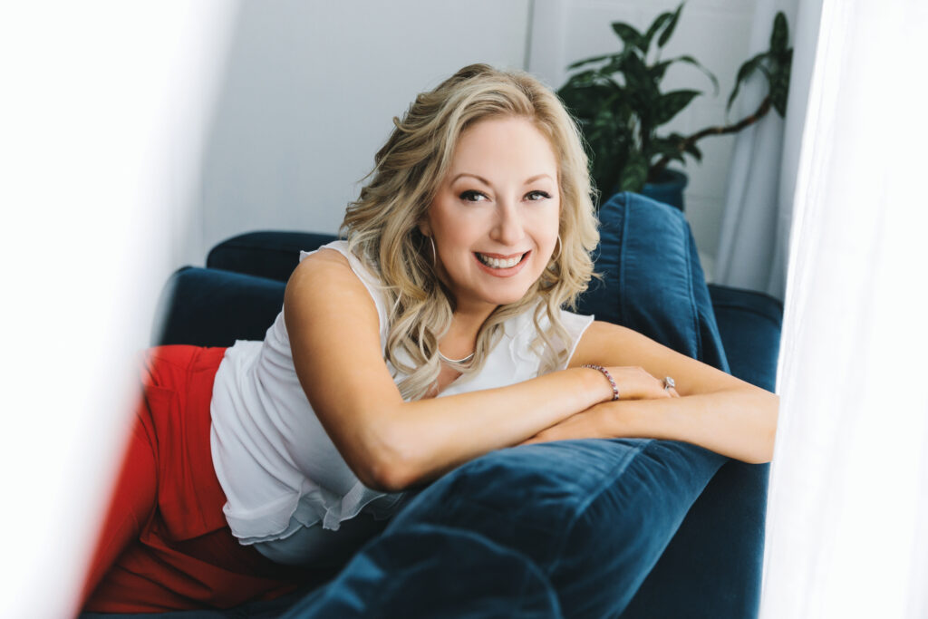 Woman in white blouse and red pants for a branding photography session sitting on a blue couch by Lindsay Hite