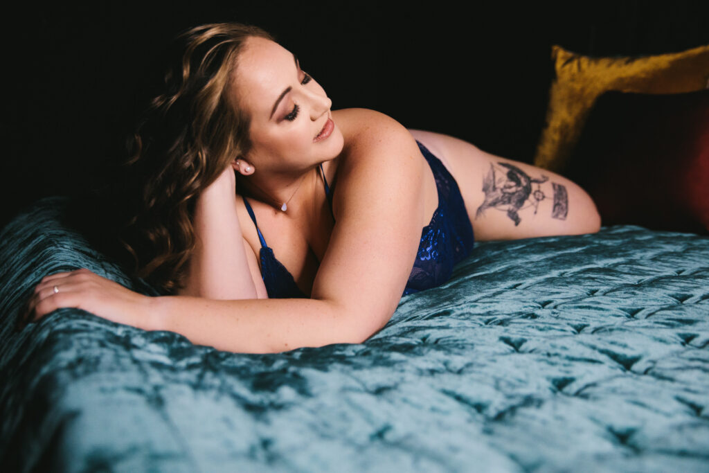 Woman in navy blue lingerie leaning on her forearm on a jewel-toned bed. Photography by Lindsay Hite. 