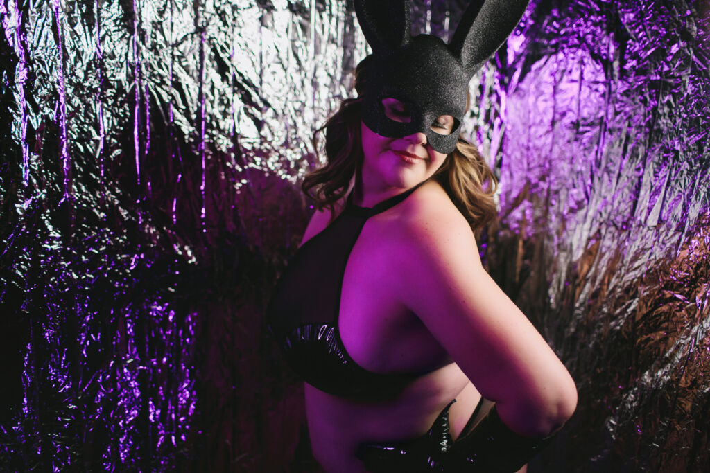 Portrait of a woman in leather lingerie and a black bunny mask in front of a psychedelic looking backdrop. Photography by Lindsay Hite. 