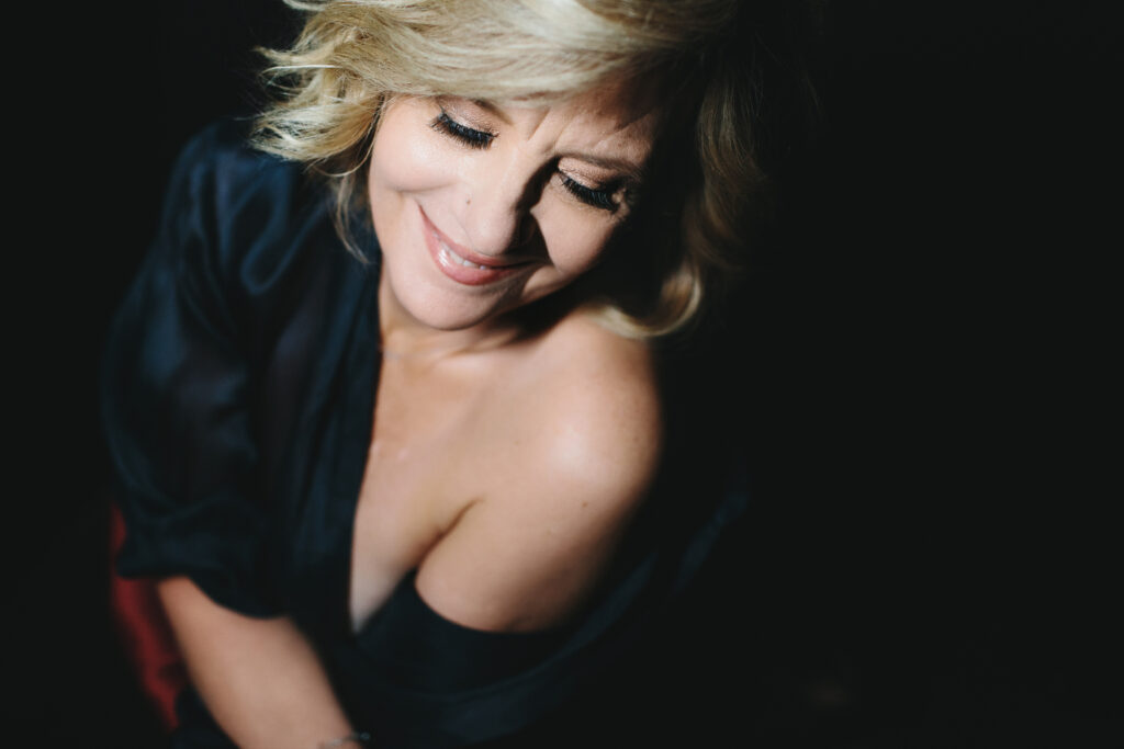 Woman with a dark robe dripping off her left shoulder with a black backdrop.  She smiles as she embraces her fierce after forty journey.  Photography by Lindsay Hite