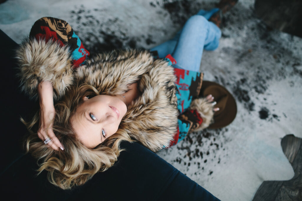 Woman wearing a fur-lined colorful jacket with jeans leaning back against a sofa.  Photography by Lindsay Hite. 