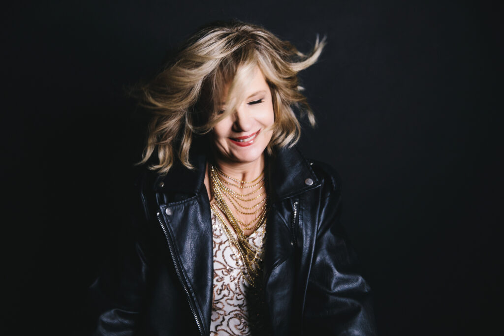 60 year old woman redefining aging and sharing her fierce after forty journey.  She's wearing a while and gold top with a black leather jacket.  There is a sense of movement in the photography with her hair and a smile on her face.  Photography by Lindsay Hite. 