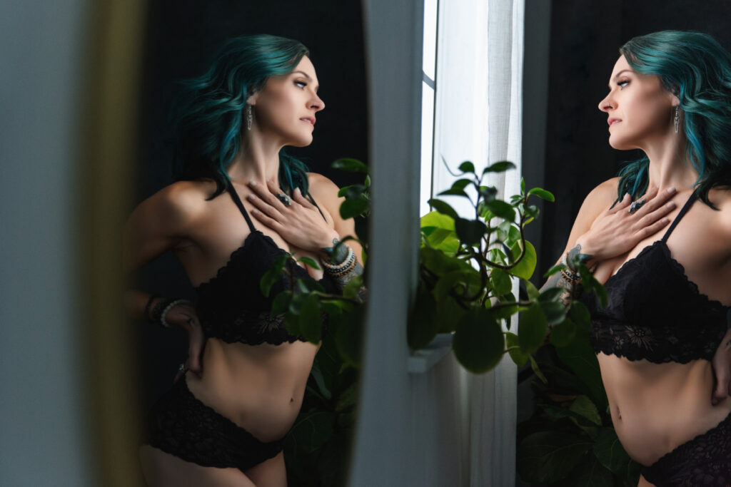 Woman with died teal hair looking out a window with a reflection of herself in the mirror.  Photography by Lindsay Hite