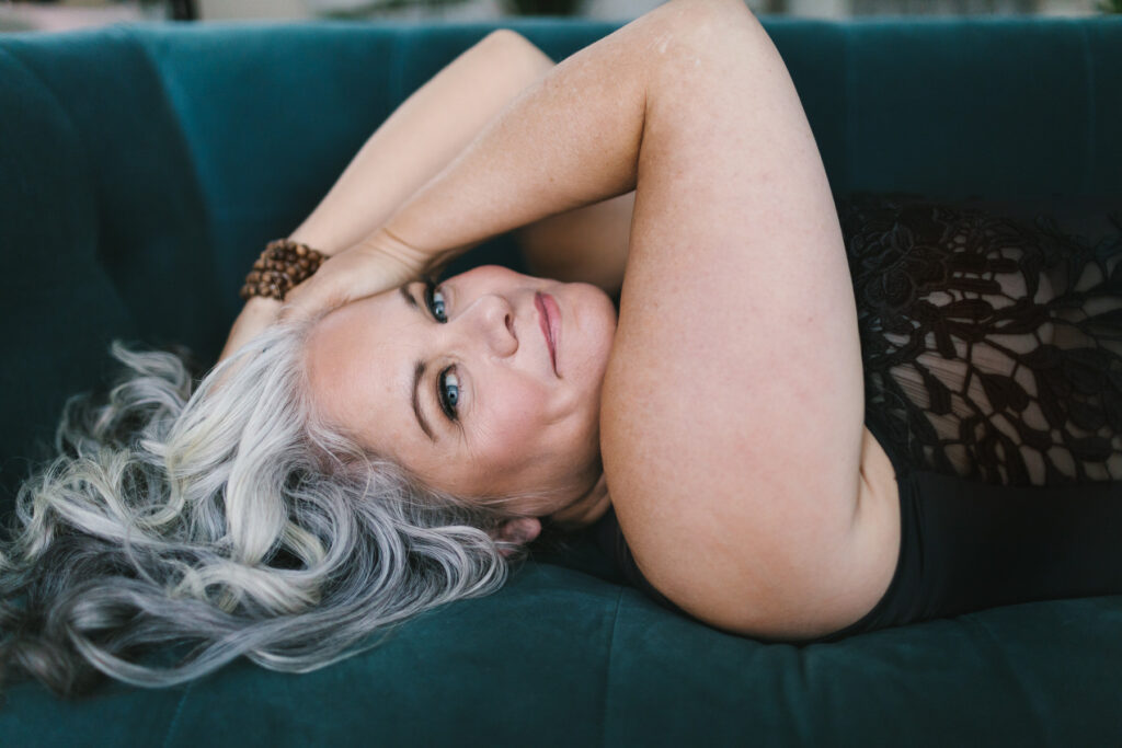 Grey haired woman in black top laying on a teal sofa smiling at the camera. Photography by Lindsay Hite